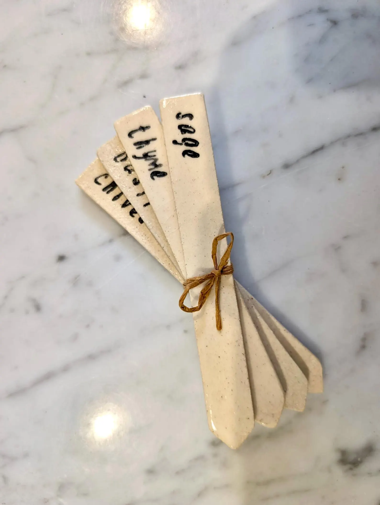 A small bundle of cream-colored ceramic recipe cards tied with a thin brown string, resting on a white marble surface. The cards are handwritten with ingredients in black ink.