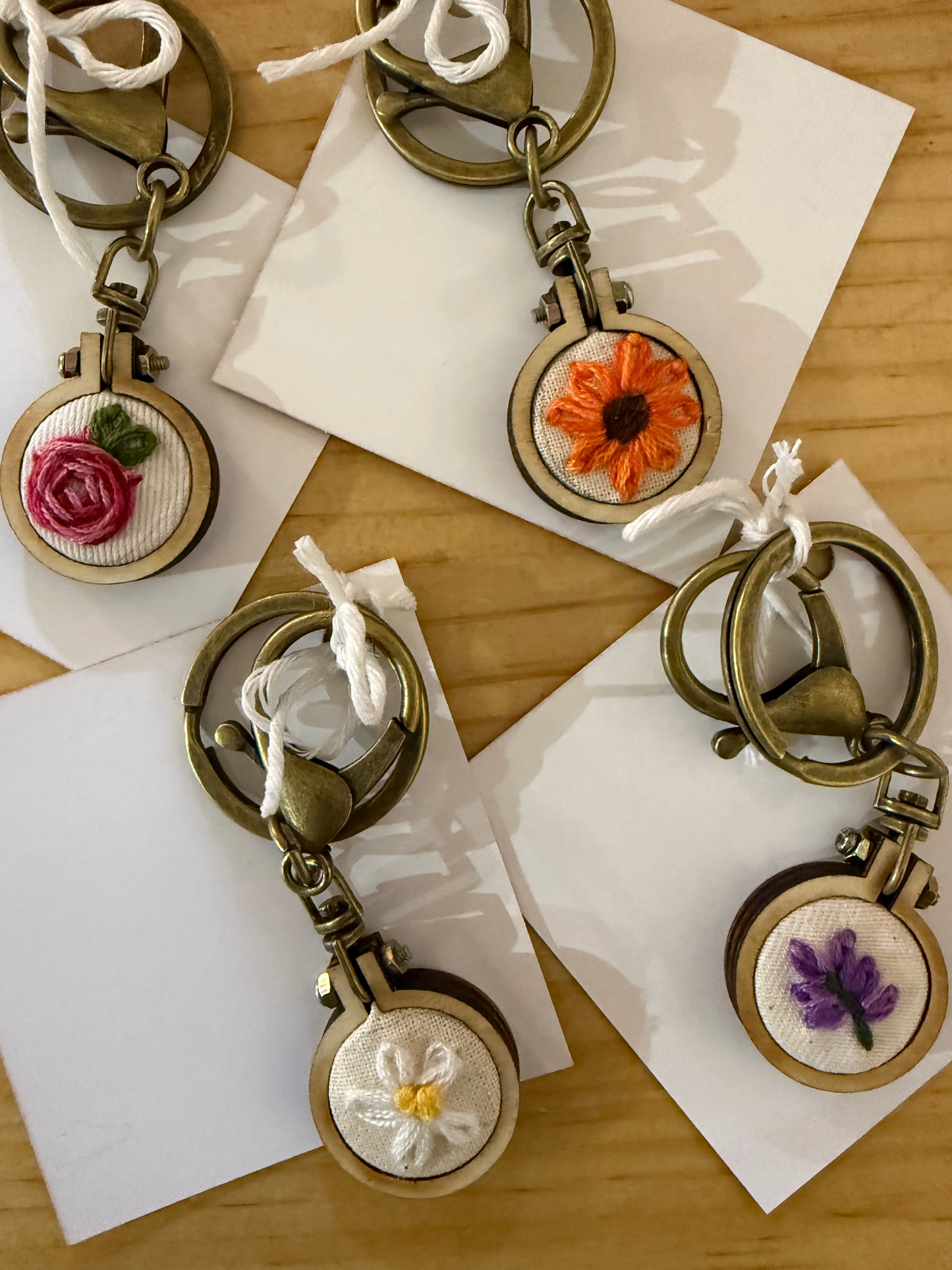 Four embroidered metal keychains with antique bronze-toned rings and round wooden medallions are arranged on white cards over a light wood surface. The embroidery features pink rose, orange flower, white daisy, and purple lavender designs.