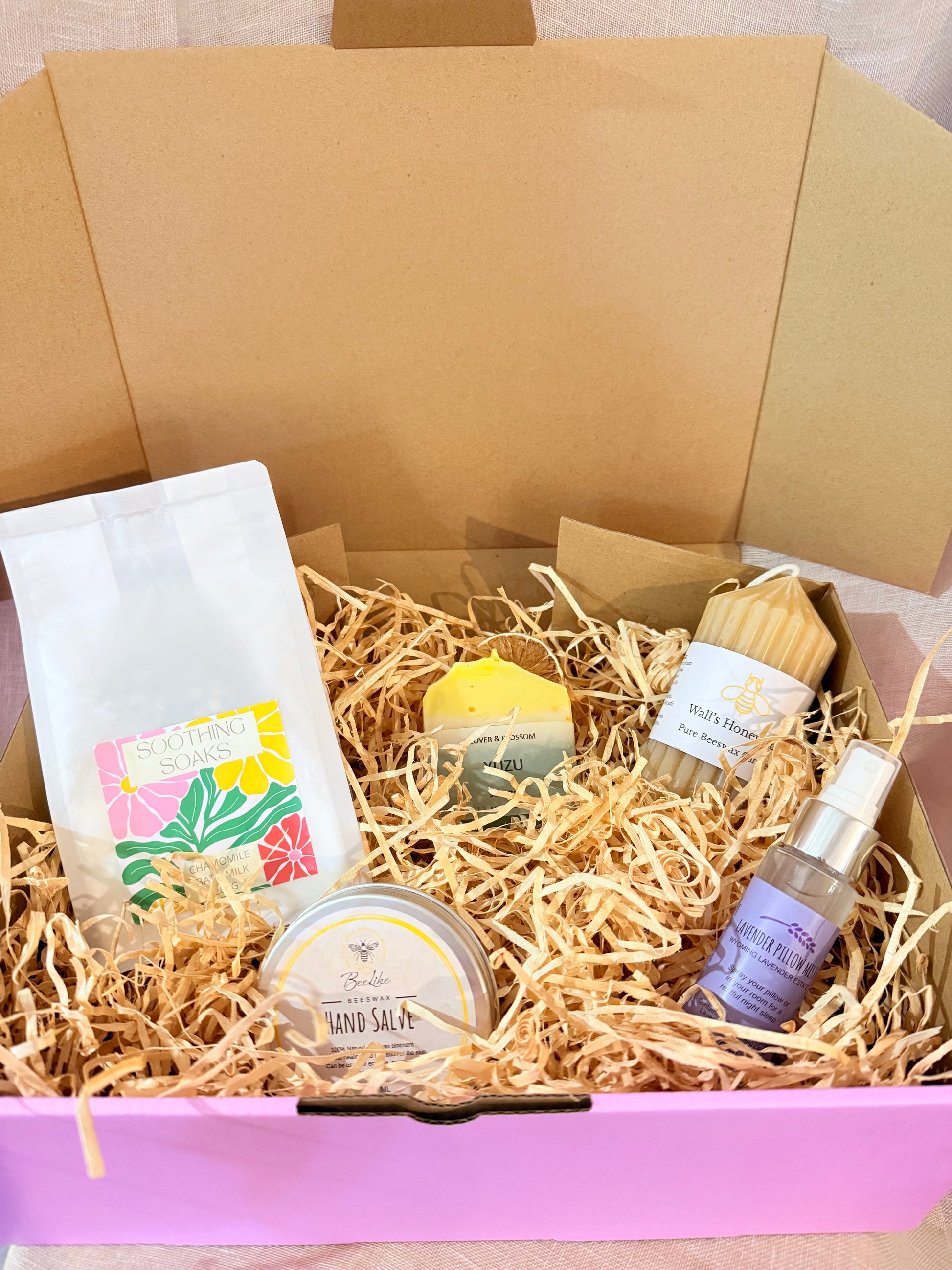 Box of bath and body products including a bag, soap, and other items on a neutral background
