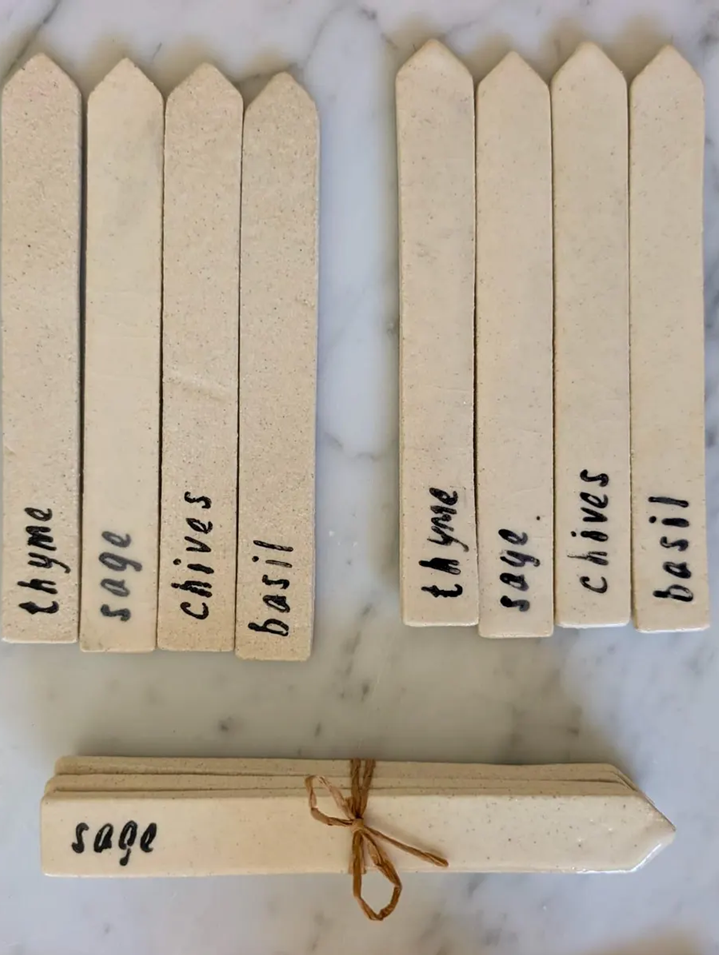 Natural wood herb labels on a white marble surface, including individual sticks labeled thyme, sage, chives, basil, and a small bundle tied with twine also labeled sage.