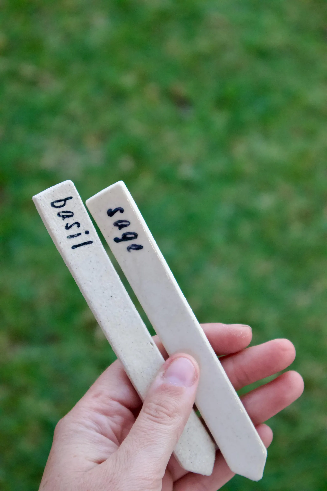 A hand holding two light beige ceramic plant labels with handwritten names 'basil' and 'sage' against a blurred green grass background.